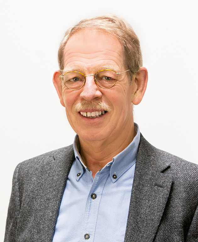 Ein älterer Mann mit hellem Haar, Brille, blauem Button-Down-Hemd und grauem Blazer lächelt vor einem schlichten hellen Hintergrund in die Kamera.